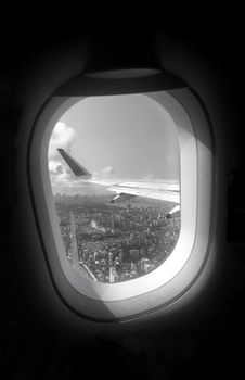Black and white aerial view through airplane window showing cityscape below with wing in view.