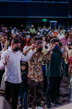 A diverse group of masked individuals holding hands and praying at an indoor gathering.