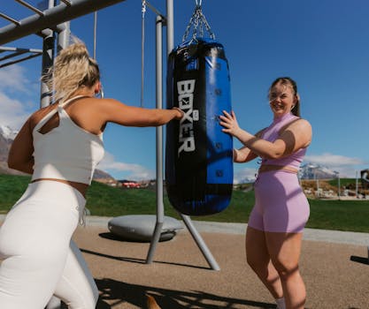 Two women exercising with a punching bag at an outdoor park, enhancing fitness and strength.