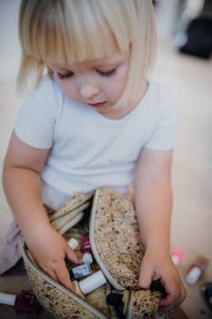 A young girl with blonde hair explores a floral cosmetic bag indoors, surrounded by beauty products.