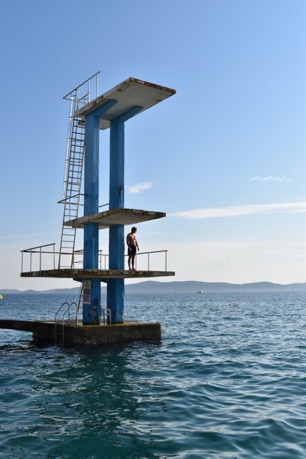 A man stands on a tall diving platform above the ocean under a clear blue sky, offering a scenic view.
