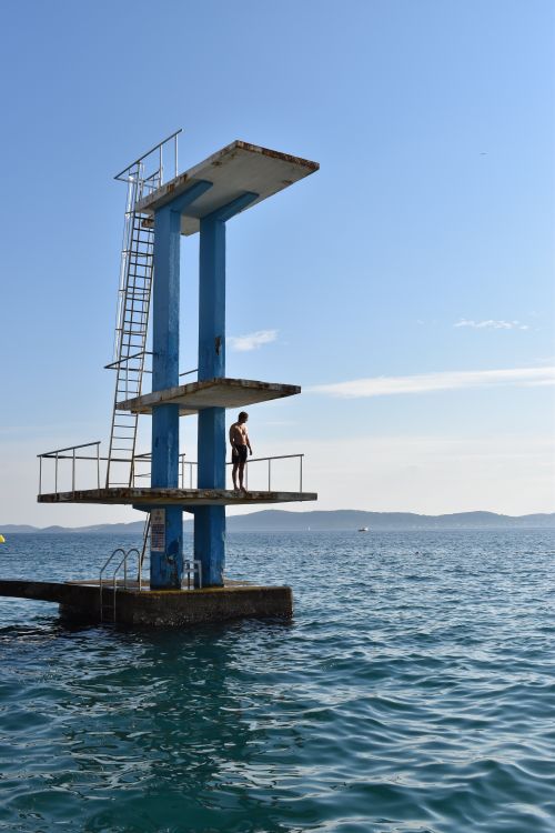 Free A man stands on a tall diving platform above the ocean under a clear blue sky, offering a scenic view. Stock Photo