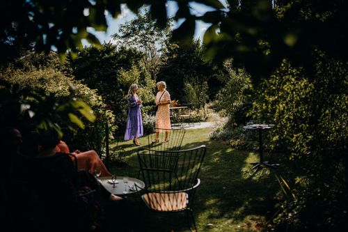 Free Two women engage in conversation in a lush summer garden setting, surrounded by chairs and tables. Stock Photo