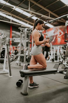 Woman kneeling on a bench exercising with dumbbells in a modern gym setting.