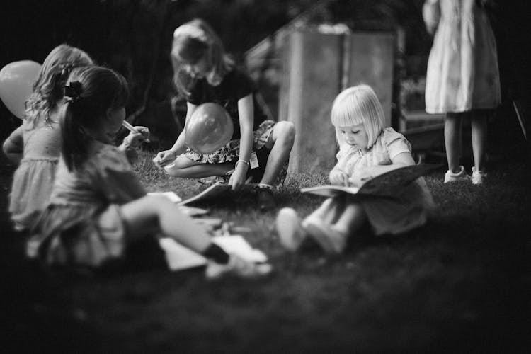 Grayscale Photo Of Kids Reading Books While Sitting On The Grass Field 