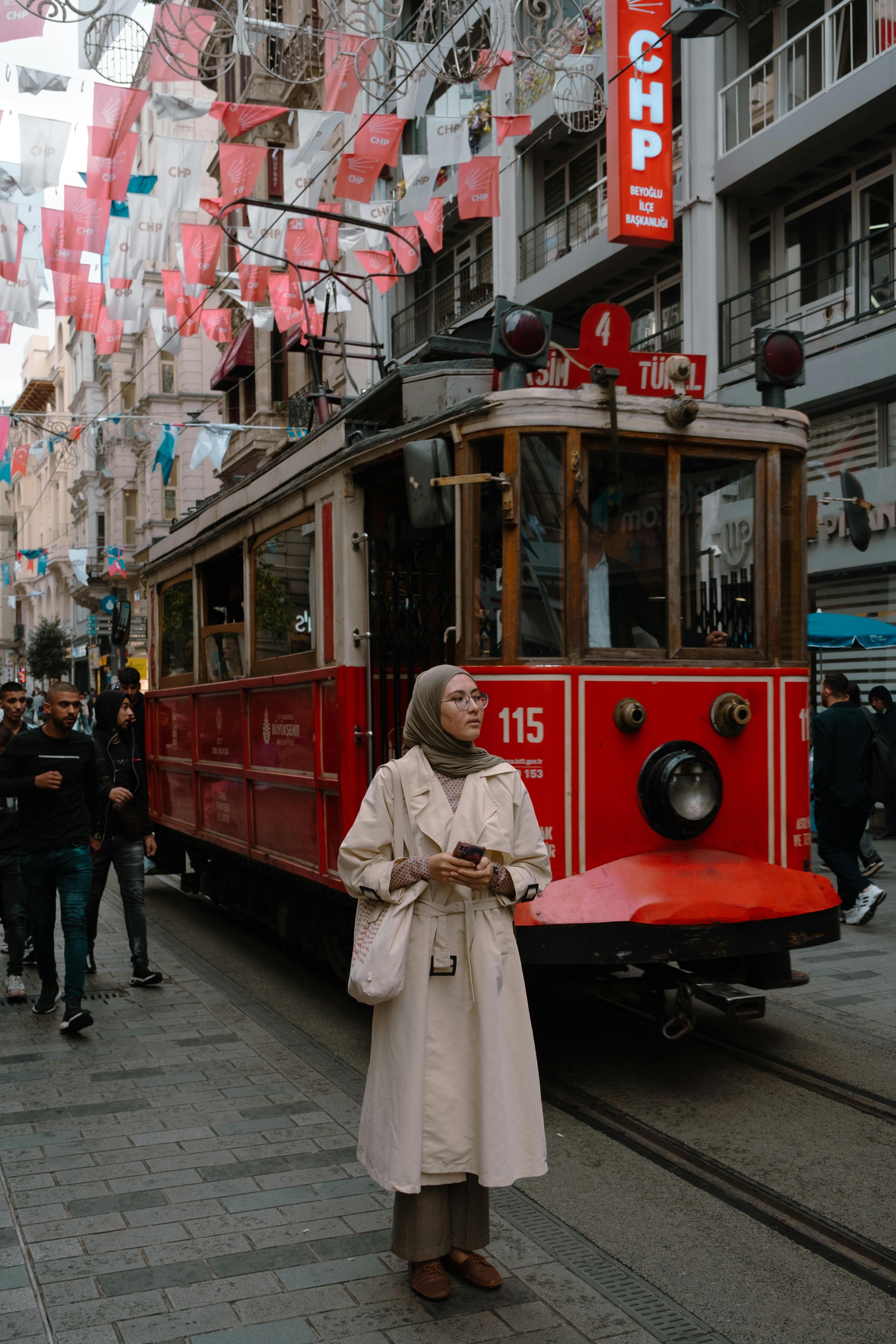 A woman in a beige coat stands by a vintage red tram in a bustling city street, surrounded by vibrant flags.