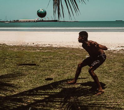 A shirtless man enjoys playing soccer on a sunny beach by the ocean.