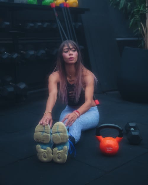 Woman in Blue Leggings Sitting on Floor Beside Kettle Bell and Dumbbell 