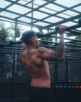 Muscular tattooed man doing pull-ups on metal bars outdoors, showcasing strength and fitness.