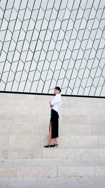 Woman in black skirt and white shirt poses on modern staircase with geometric wall.