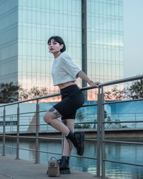 Fashionable tattooed woman in crop top and leather boots posing at urban waterfront during the day.