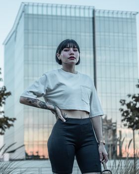 A woman poses confidently with tattoos, short hair, and stylish urban attire in front of a glass building.