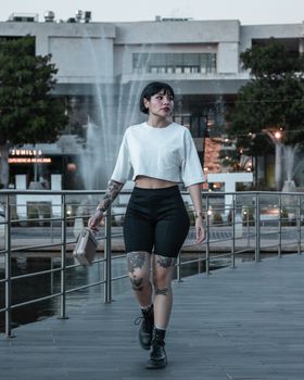 Tattooed woman in stylish attire walking by a fountain in an urban setting.