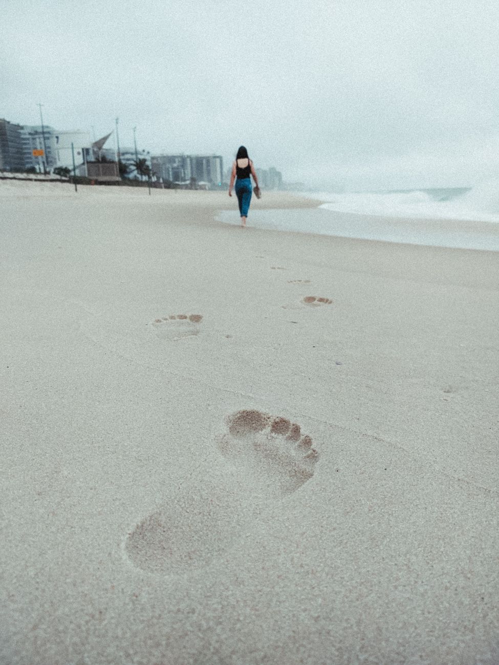 Footprints leading towards the ocean as a woman walks alone on a cloudy beach.