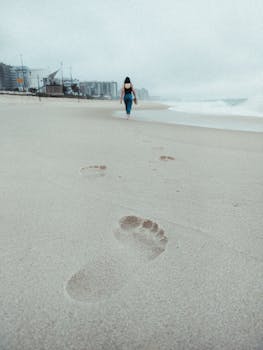 Footprints leading towards the ocean as a woman walks alone on a cloudy beach.