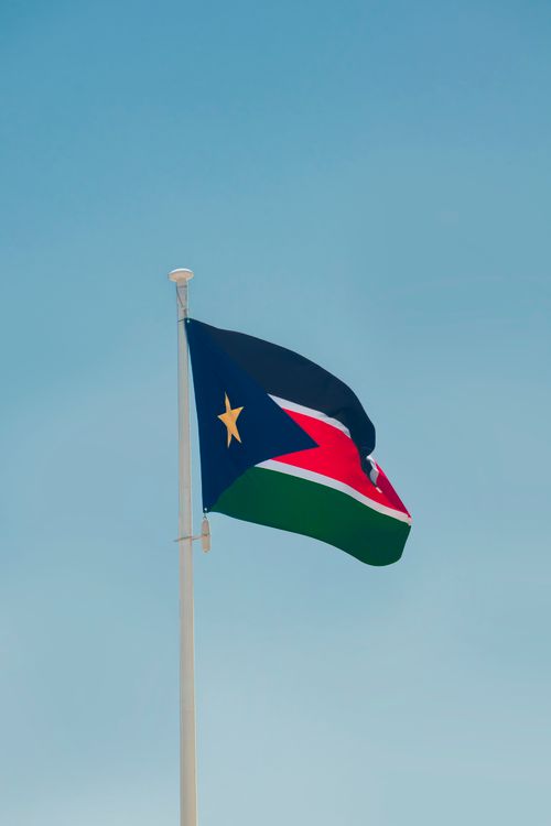 Free South Sudan national flag fluttering on a flagpole under a clear blue sky, symbolizing patriotism. Stock Photo