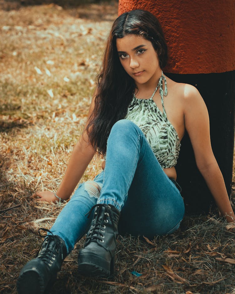 Young Woman Posing In Printed Halter Crop Top Ad Denim  Pants Sitting On Grass