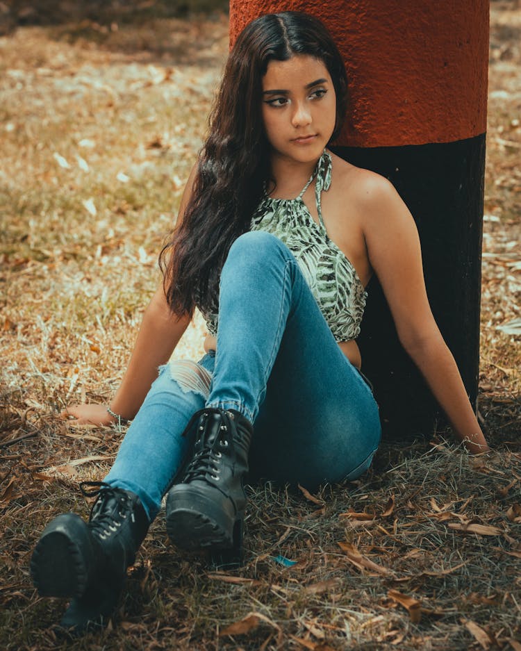 Young Woman Posing In Printed Halter Crop Top Ad Denim Pants Sitting And Leaning On A Painted Concrete Post