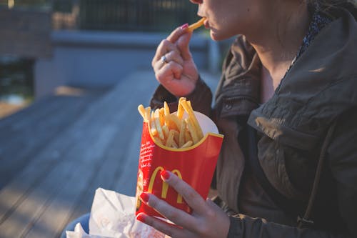 Woman in Brown Classic Trench Coat Eating Mcdo Fries during Daytime