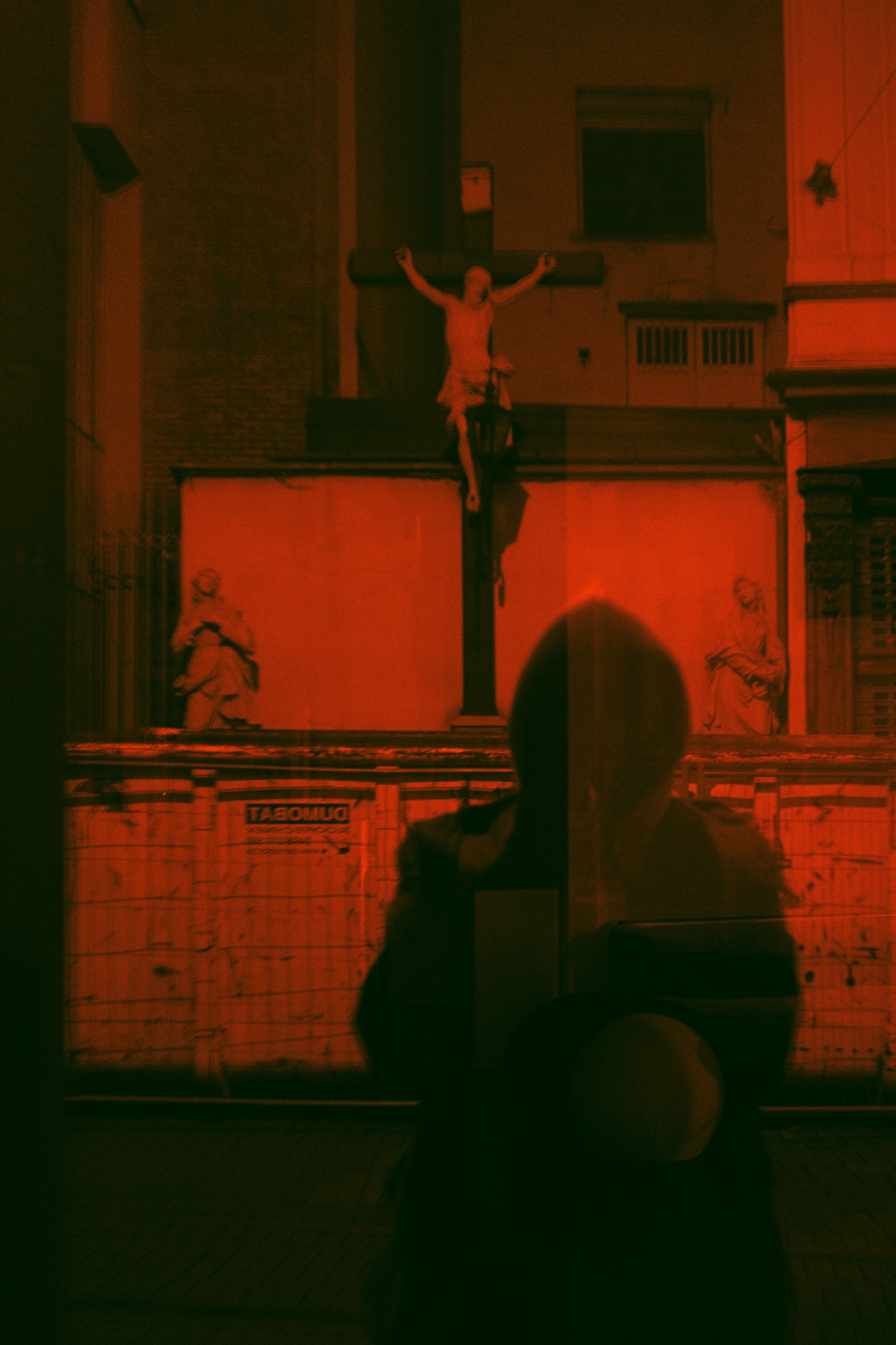 A silhouette reflects on a glass with a cross and statue in an urban backdrop, enveloped in dark orange hues.