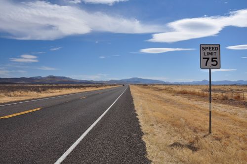 Free A deserted highway stretches into the horizon under a bright blue sky with a speed limit sign in the countryside. Stock Photo