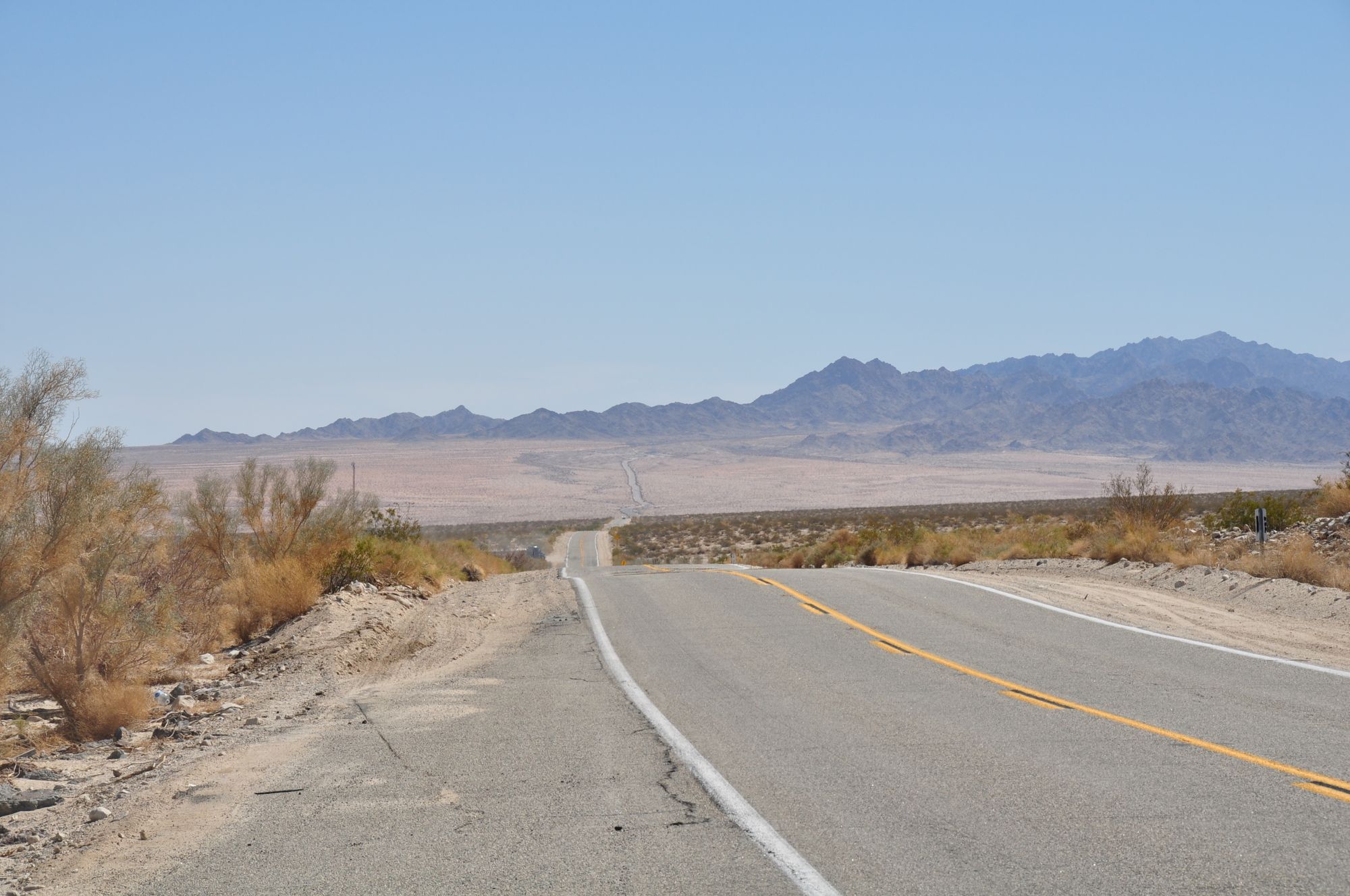 A solitary asphalt road stretches across the arid Californian desert, surrounded by mountains and clear blue skies.
