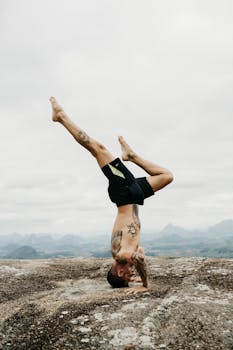 Shirtless man balancing in a headstand with tattoos, set against a foggy mountainous backdrop. Fitness and nature combine.