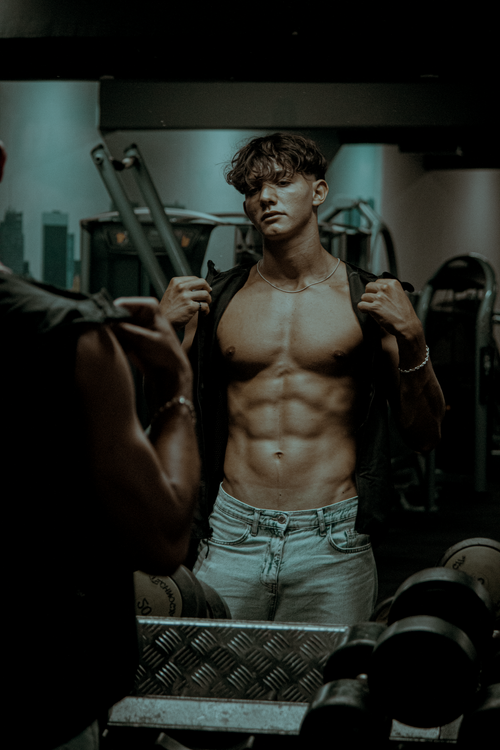 Free Muscular young man showing his abs in a gym mirror, highlighting strength and fitness. Stock Photo