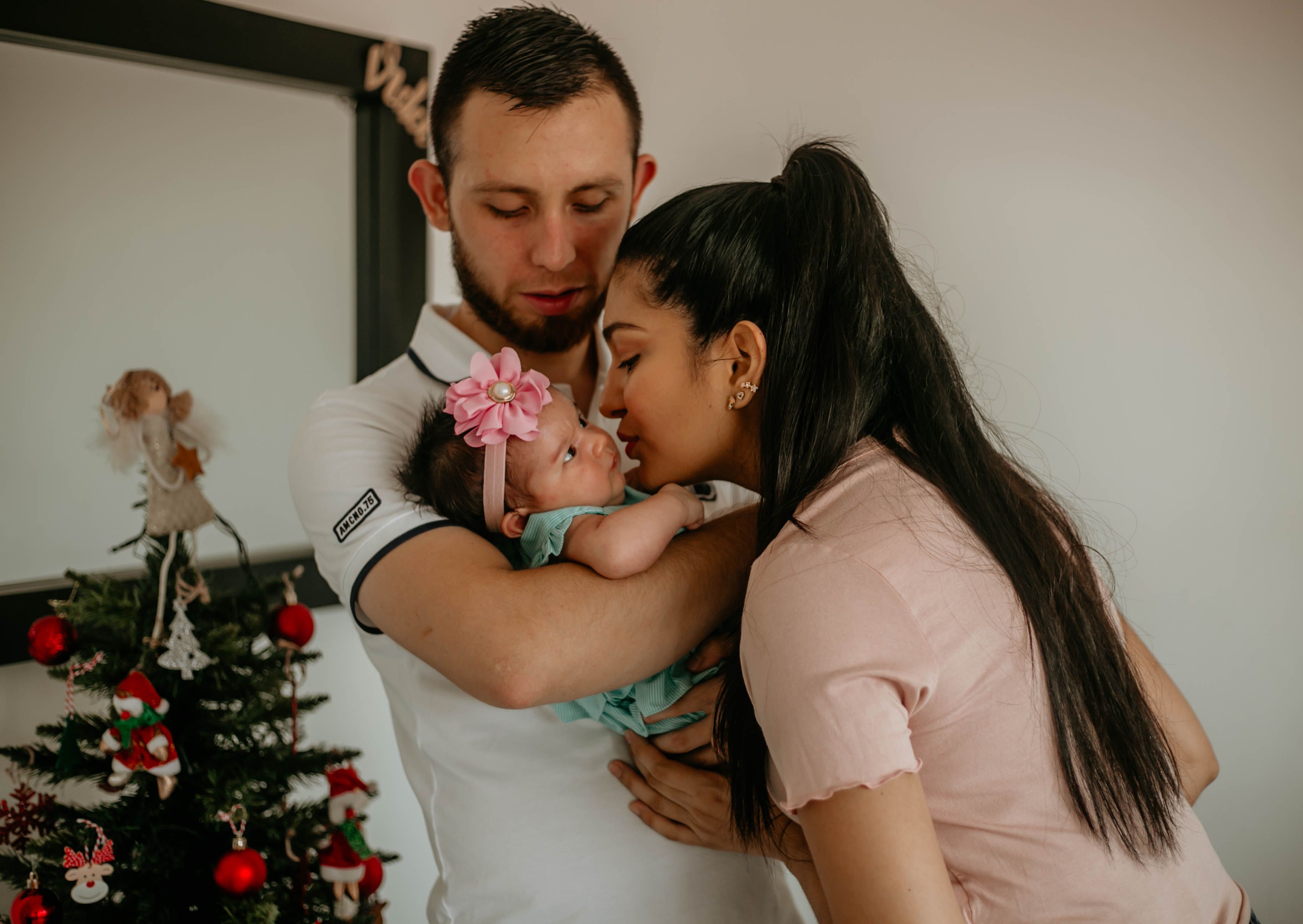 A loving couple kisses their baby beside a decorated Christmas tree.