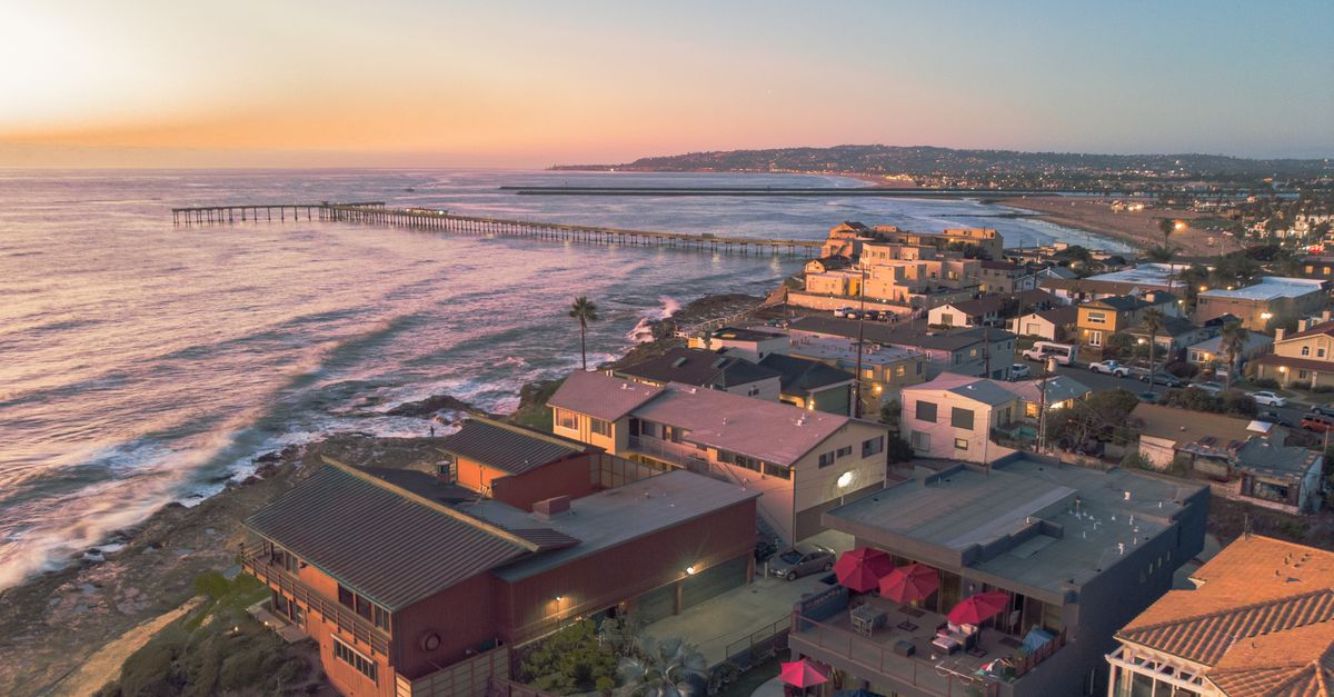 Stunning aerial shot of coastal homes and pier at sunset, waves crashing on the shore.