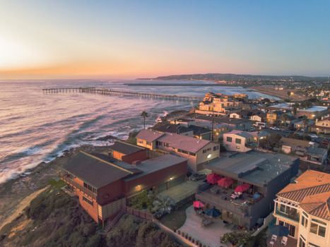 Stunning aerial shot of coastal homes and pier at sunset, waves crashing on the shore.