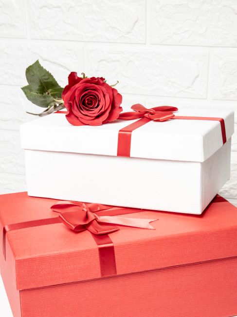 Fotomural Una caja de regalo blanca con una cinta roja y una rosa encima de una caja roja contra una pared texturizada blanca.