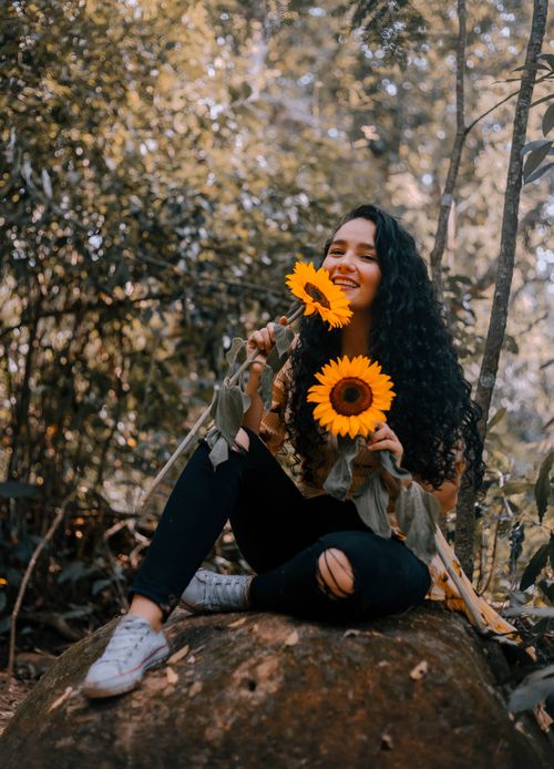 Free Joyful woman sitting on a rock, holding vibrant sunflowers amidst greenery. Stock Photo