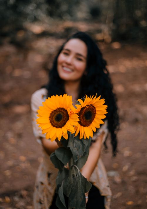Free A smiling woman with long curly hair holds vibrant sunflowers in an outdoor setting, symbolizing joy. Stock Photo