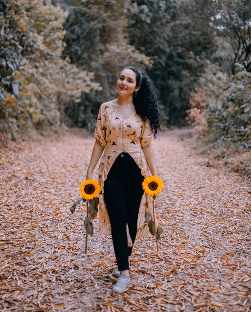 Free A smiling woman walks on a leaf-covered path holding sunflowers, embracing autumn's beauty. Stock Photo