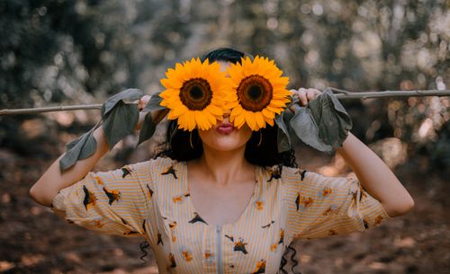 Free A playful woman in a floral dress holds sunflowers over her eyes in a vibrant outdoor setting. Stock Photo