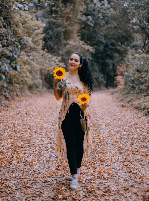 Free A woman with curly hair walks through a fall scene holding sunflowers, creating a vibrant and joyful mood. Stock Photo