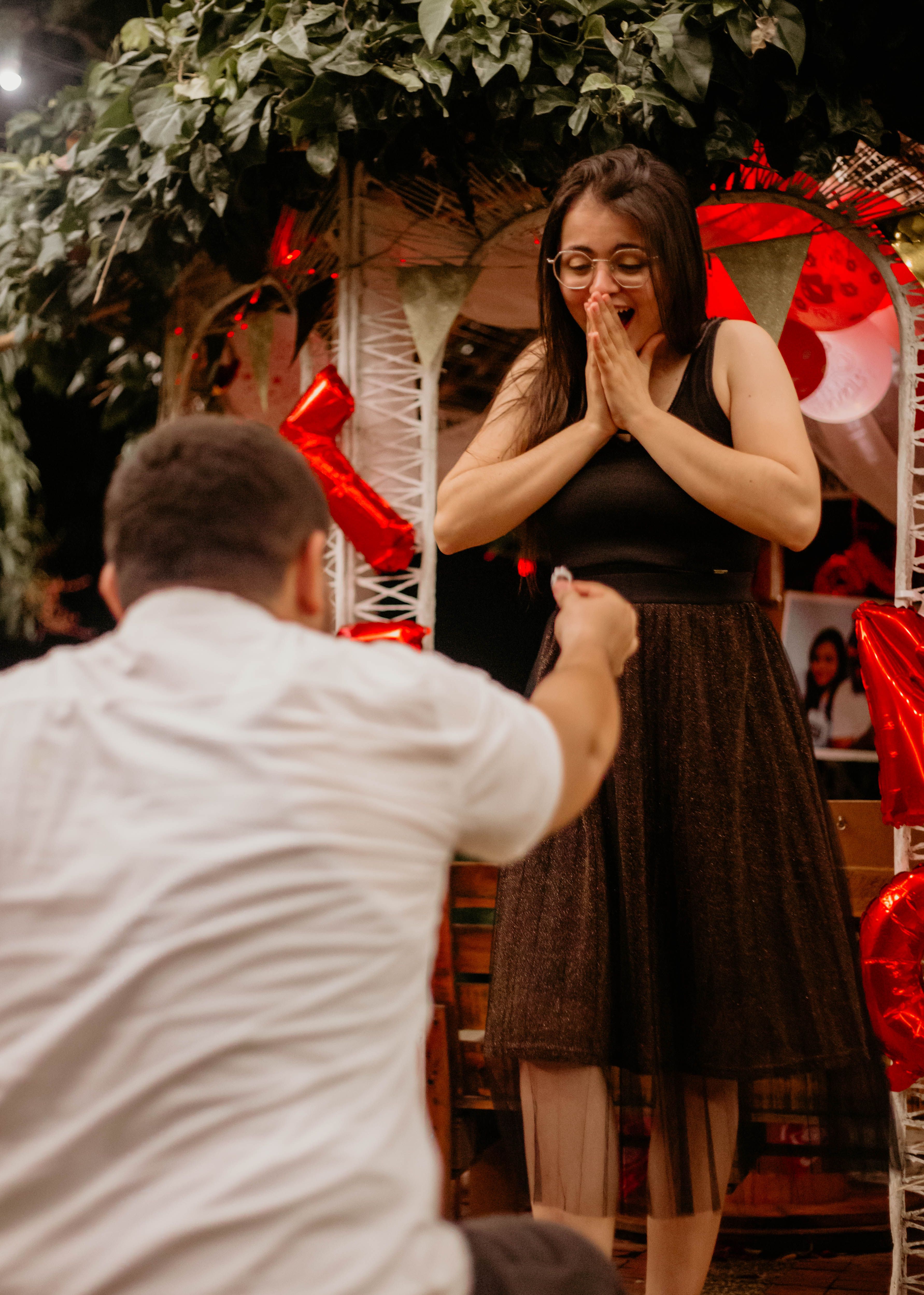 Romantic proposal scene with a man proposing to a surprised woman under lush greenery and red heart decorations.