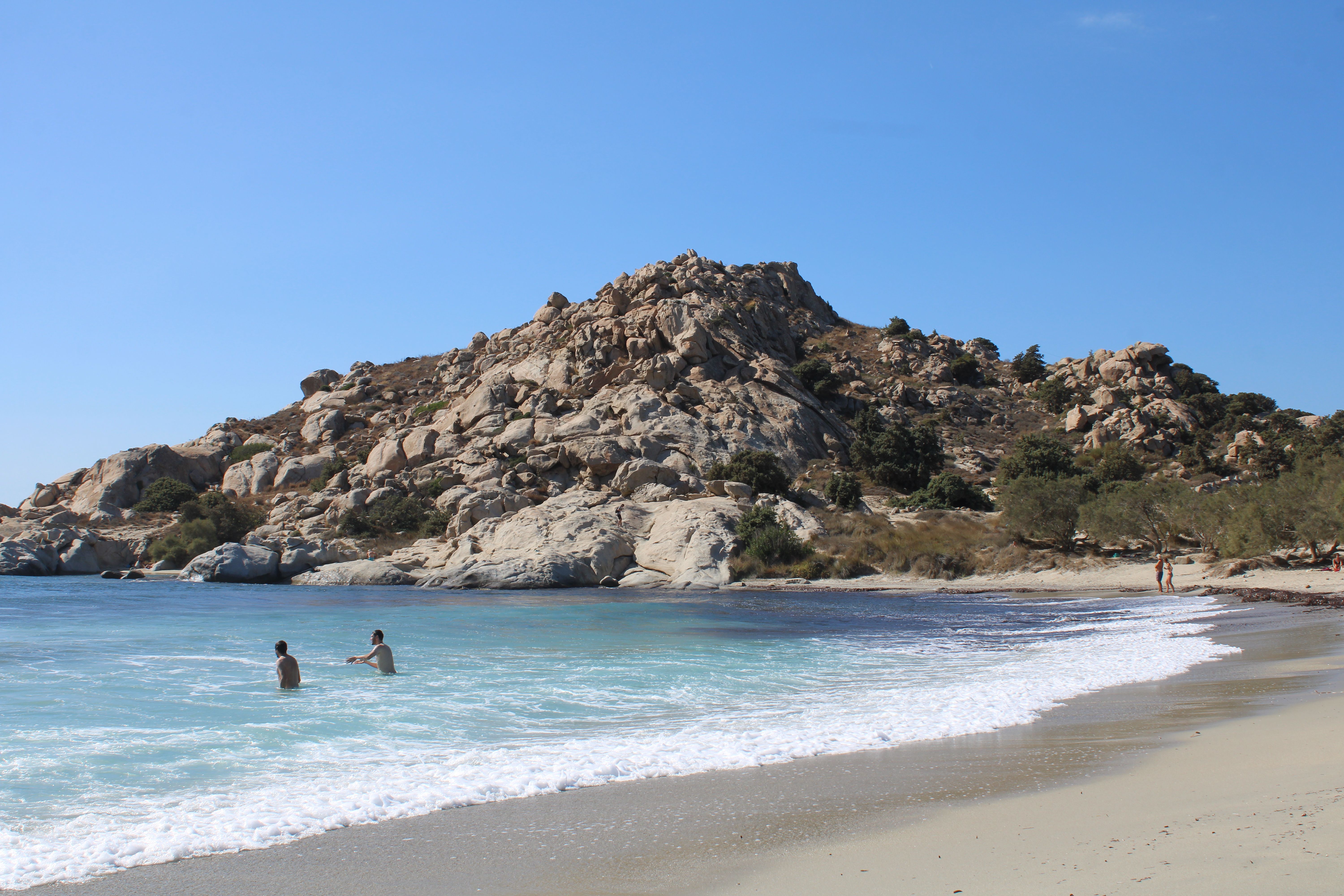 Tourists enjoying a sunny day at Mikri Vigla beach with rocky hills and clear blue water.