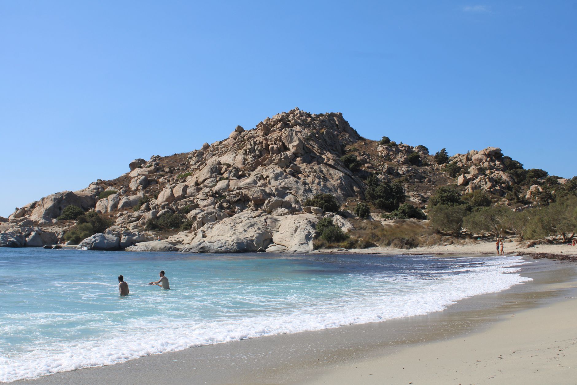 Tourists enjoying a sunny day at Mikri Vigla beach with rocky hills and clear blue water.