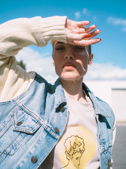 Fashionable woman posing outdoors on a sunny day, shielding eyes from the sun with denim jacket and casual attire.