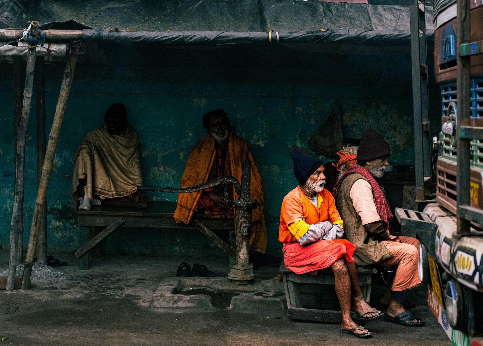 Elderly men sitting by a traditional water pump on an urban street, dressed in vibrant clothing.