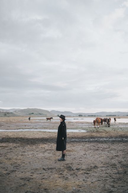 A woman in a hat stands in a vast, overcast field with grazing horses.