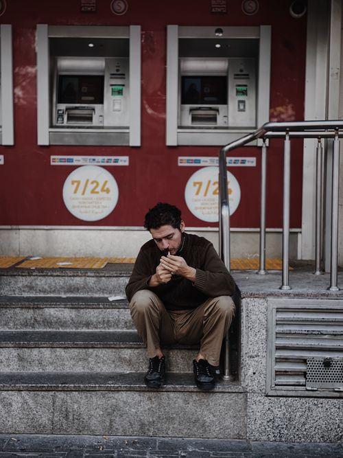 Free A man sits on steps smoking near ATM machines, reflecting urban lifestyle. Stock Photo