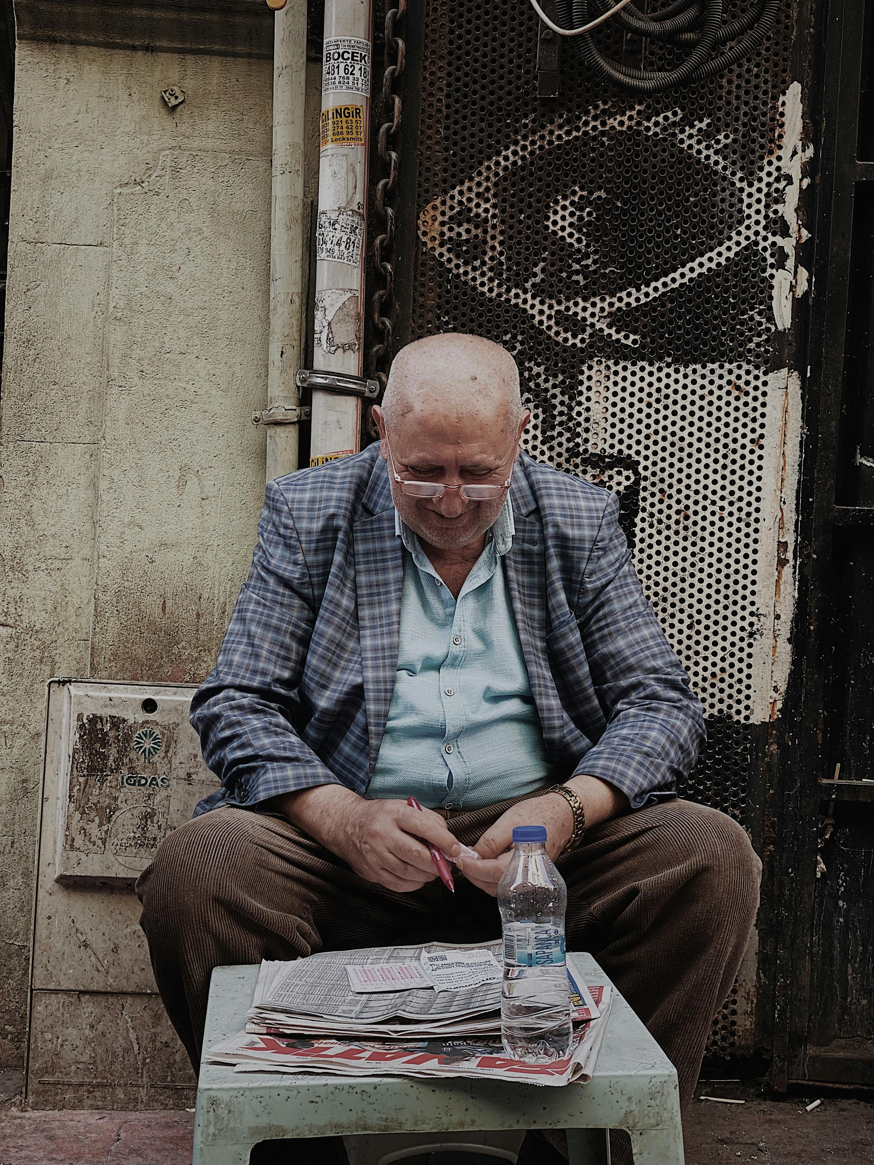 Senior man in plaid blazer sitting and reading a newspaper on an urban street.