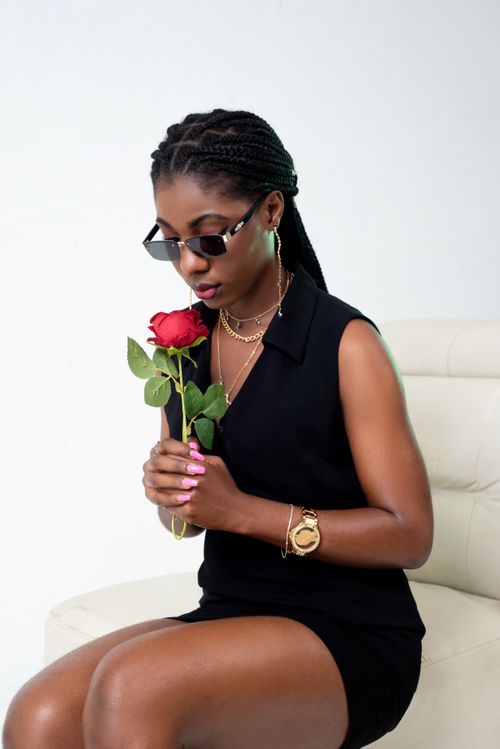 Free Stylish woman in black dress sitting indoors, holding a red rose and wearing sunglasses. Stock Photo