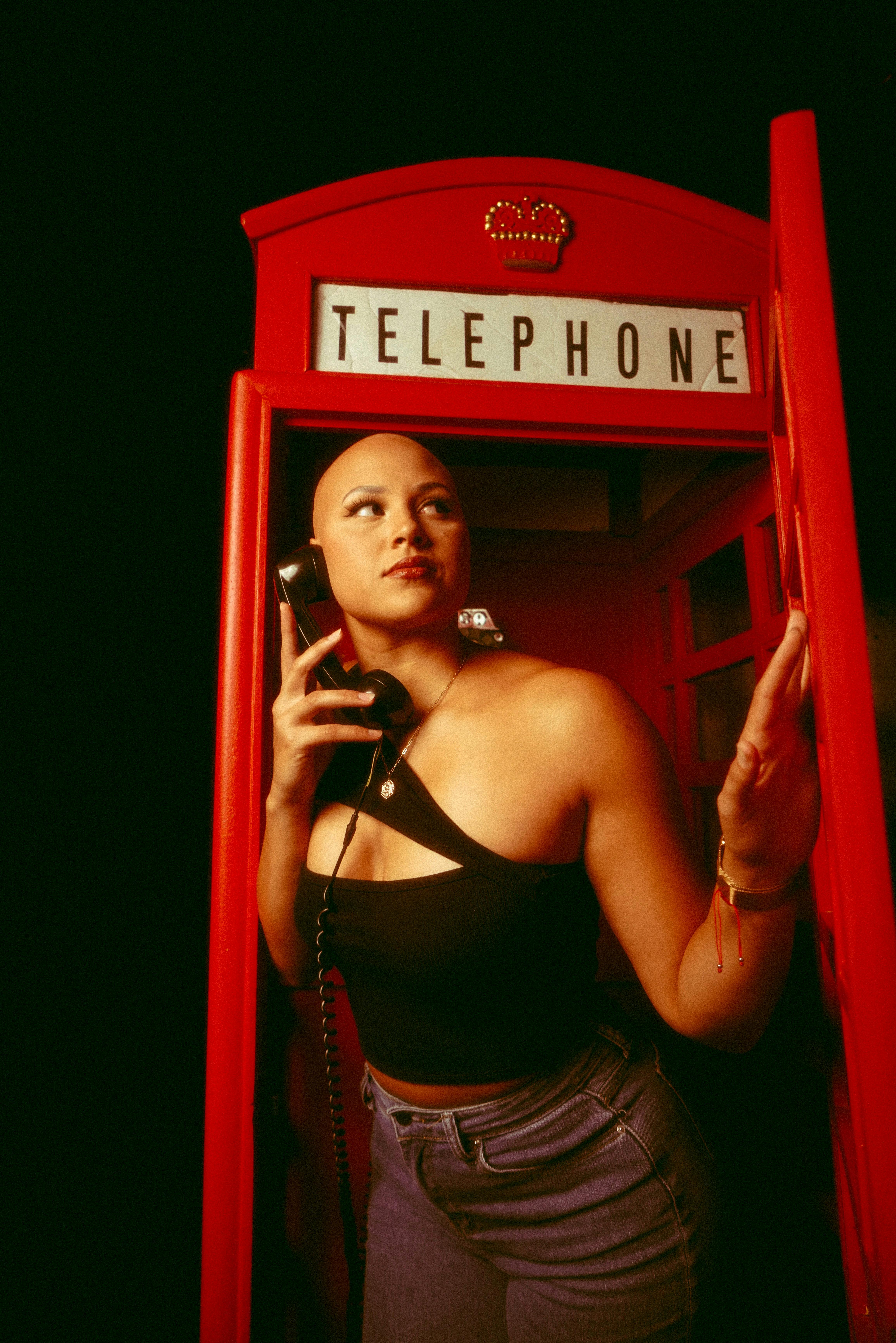 Bald woman posing confidently inside a classic red telephone booth with vintage vibes.
