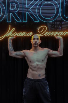 Shirtless, tattooed man flexing in gym with neon lights in the background.
