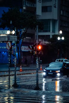 A wet city street at night featuring a red traffic light and a waiting car under rainy conditions.