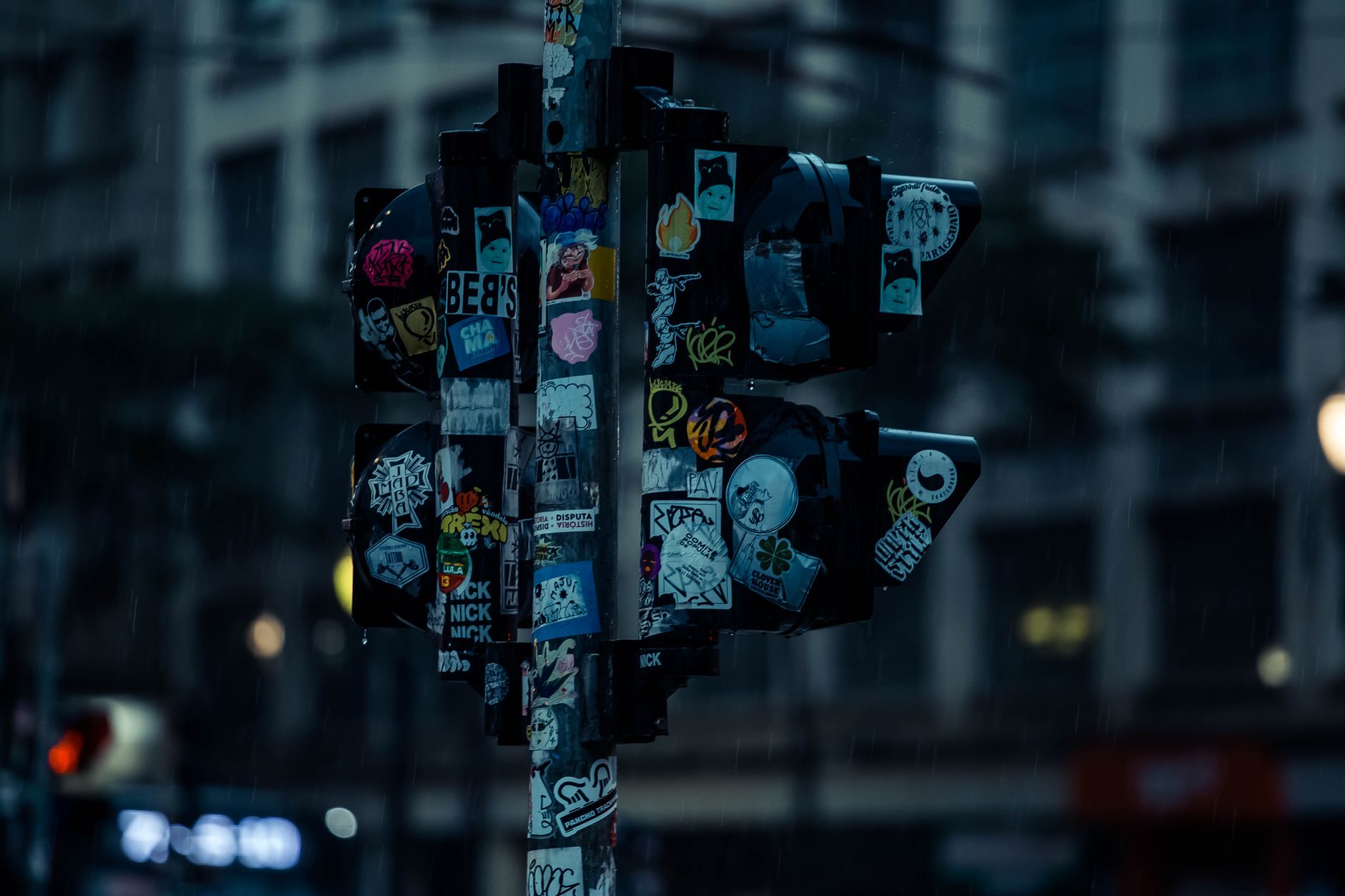 A dimly lit urban traffic light adorned with various street art stickers during a rainy night.
